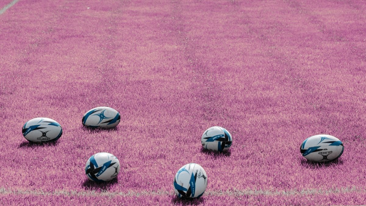 How to play women's rugby. Image shows a set of rugby balls on a field of magenta grass.
