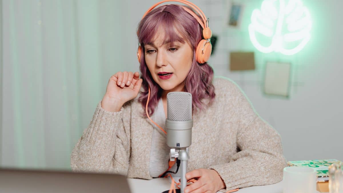 image shows a woman with pink hair speaking into a microphone as if she's recording a podcast