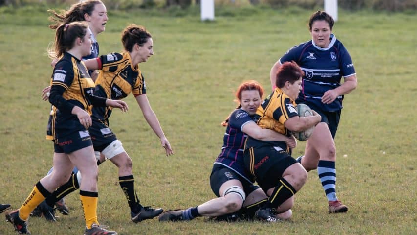 How to play women's rugby. Image shows women playing rugby with one woman tackling another during a game.